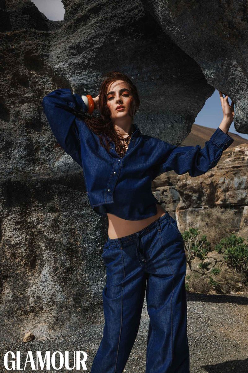 A woman in a denim outfit poses near a rock formation against a background of cliffs and blue sky, fashion photography.