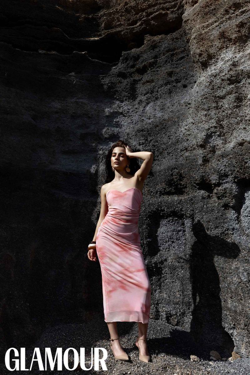 A woman in a pink dress poses in front of a dark rock formation for a fashion shot. She's wearing jewelry and high heels.