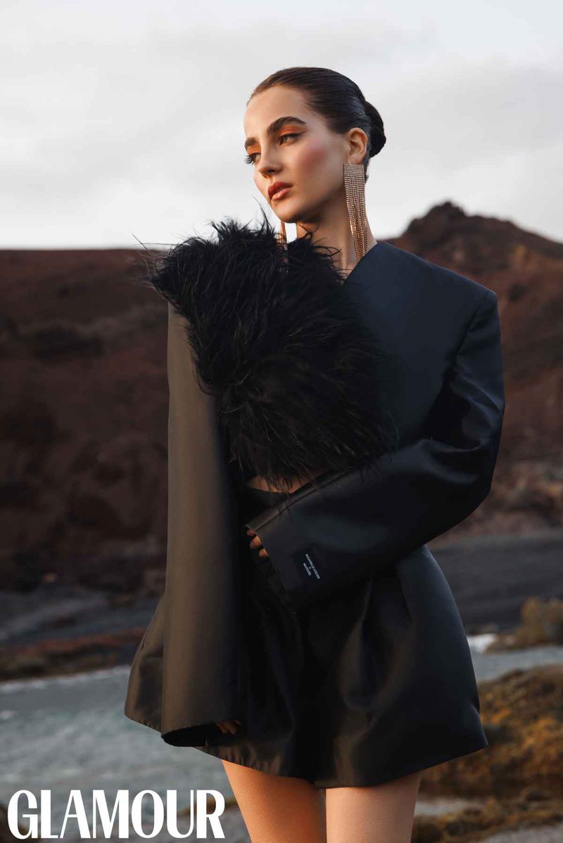 A woman in a black dress poses outdoors with feathery accessory, against a landscape of brown hills and a cloudy sky for a Glamour magazine editorial.