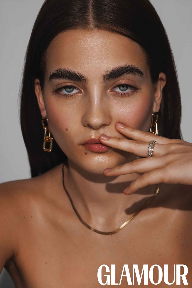 Close-up studio shot of an adult woman with medium-length brown hair wearing gold jewelry against a neutral background.
