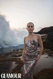 Woman in sequined floral dress standing on rocky shore with crashing ocean waves behind her at sunset