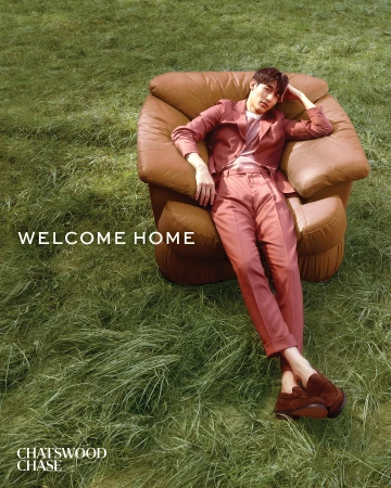 An Asian man in a pink suit reclines in a large brown leather chair, set on a grassy lawn, styled in an outdoor fashion editorial.