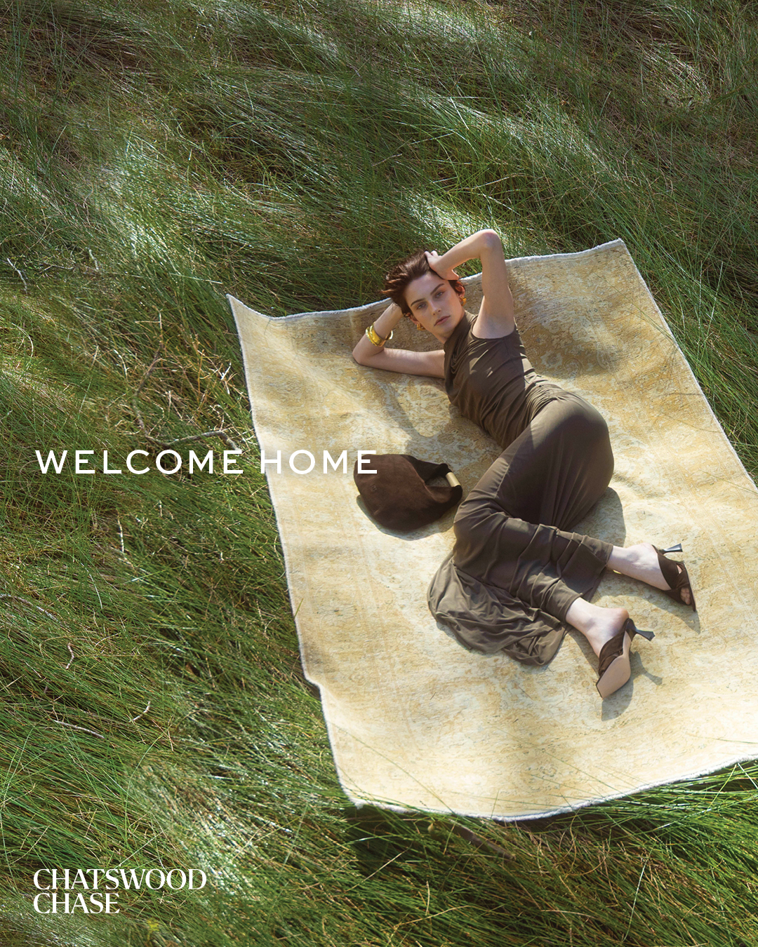 Woman in olive jumpsuit and heels lounges on a cream blanket in green field. Model poses with hands behind head, outdoors.