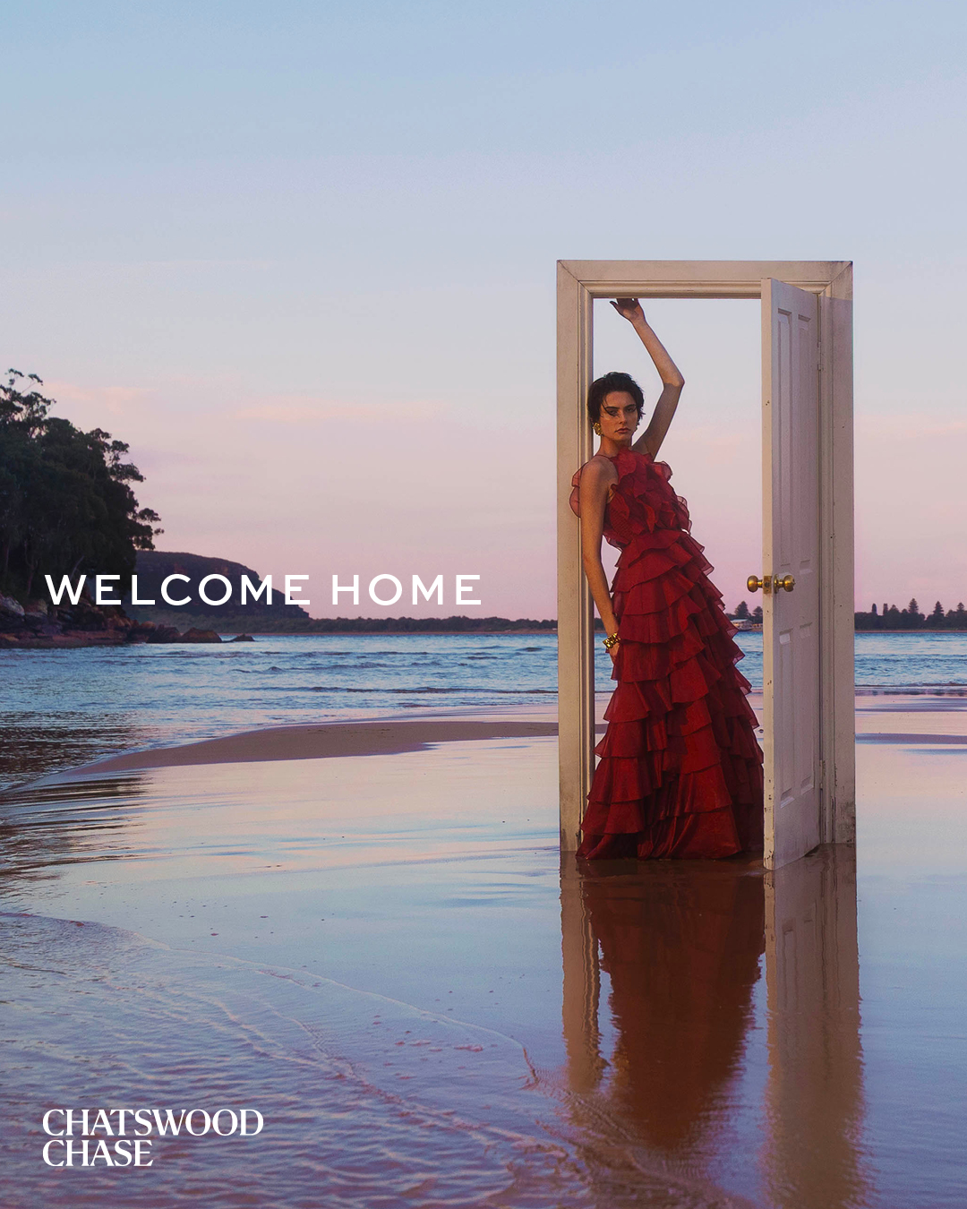 A woman in a red dress stands in a doorway on a beach at sunset, with ocean waves reflecting the sky, creating a surreal fashion scene.
