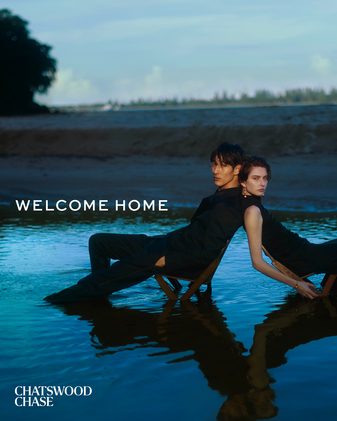 A man and a woman sit back-to-back in chairs in shallow water near the shore, captured in a cinematic, subdued lighting setting.