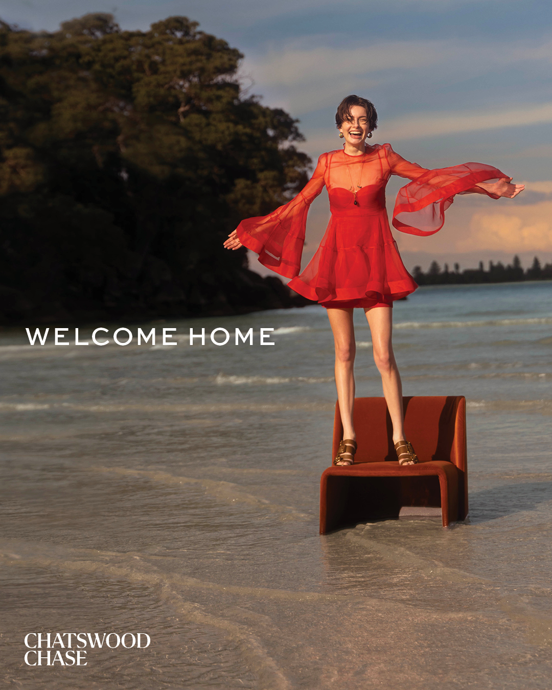 Woman in red dress stands on chair at beach with ocean and sky background under bright sunlight, wearing casual footwear and jewelry.