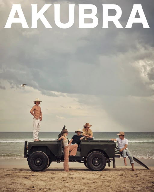 Five people with hats pose near a vintage car on a sandy beach under a cloudy sky. 'AKUBRA' text overlay.