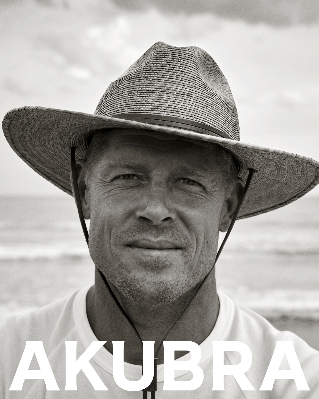 Monochrome portrait of a man wearing a hat on a beach, featuring ocean waves and the text 'AKUBRA' in bold white letters.