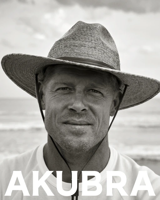 Monochrome portrait of a man wearing a hat on a beach, featuring ocean waves and the text 'AKUBRA' in bold white letters.