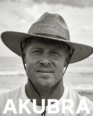 Monochrome portrait of a man wearing a hat on a beach, featuring ocean waves and the text 'AKUBRA' in bold white letters.