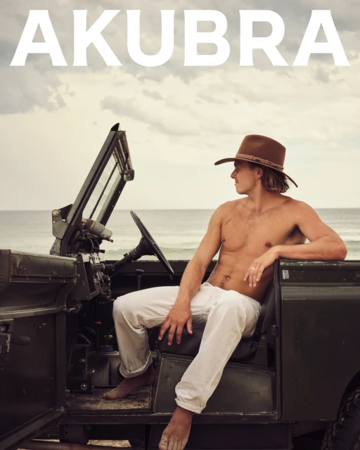 Shirtless man sits in an open jeep on a beach wearing a hat. He is looking out towards the ocean under a cloudy sky.