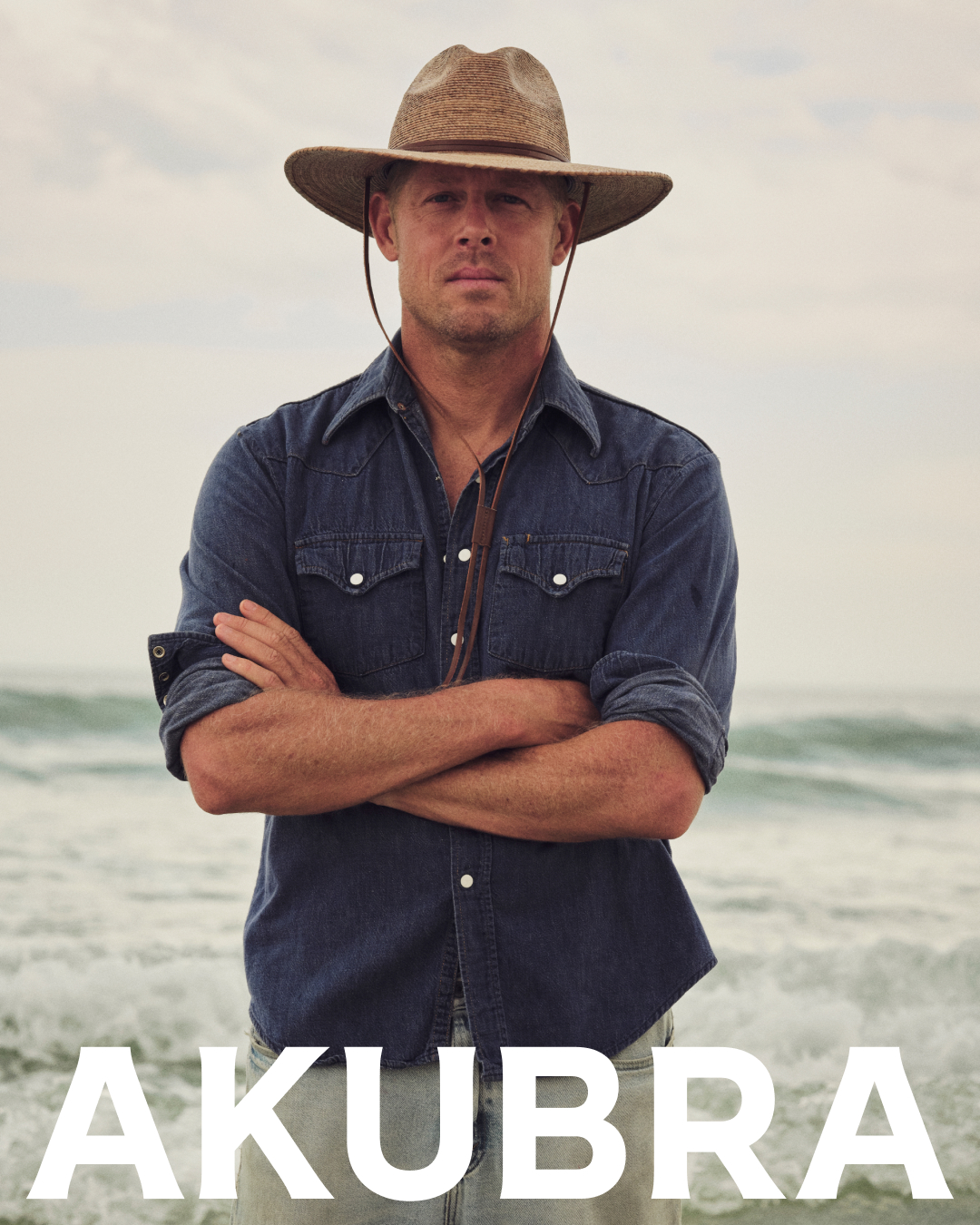 Portrait of adult male wearing a hat and denim shirt standing on a beach with a neutral expression, showcasing neutral and earth tones.