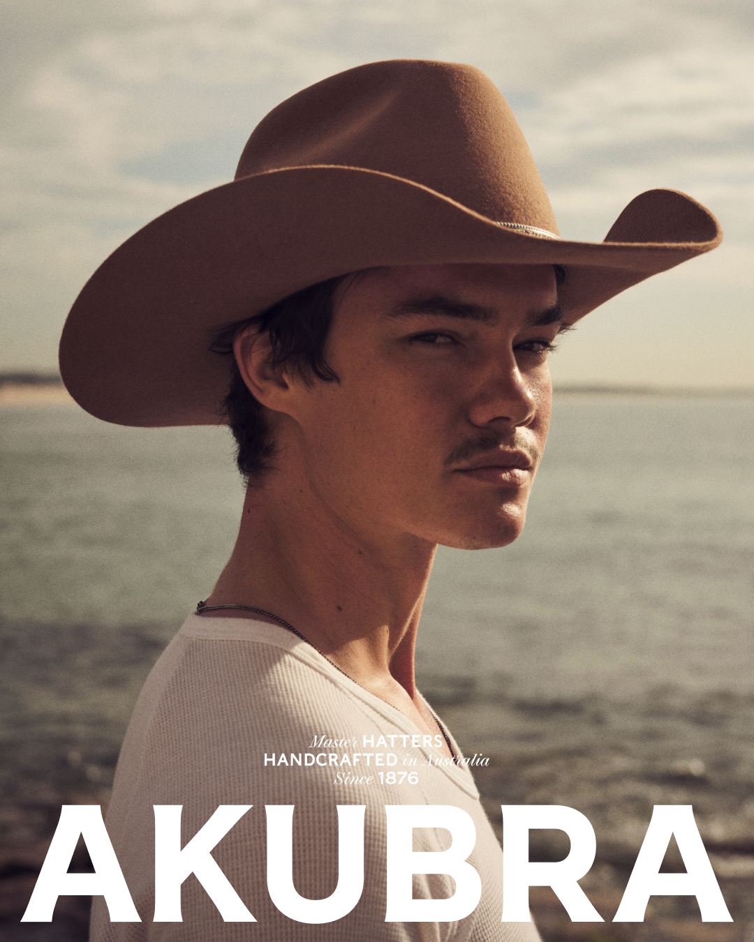 Portrait of a light-skinned adult male in a white shirt and brown cowboy hat, posing on a beach with a blurred sea background on a sunny day.