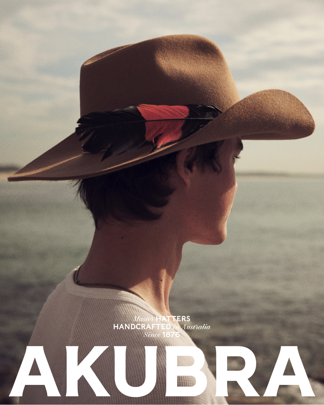 Man wearing a tan Akubra hat with a feather accent on a beach with ocean and blue sky. Akubra brand name prominently displayed.