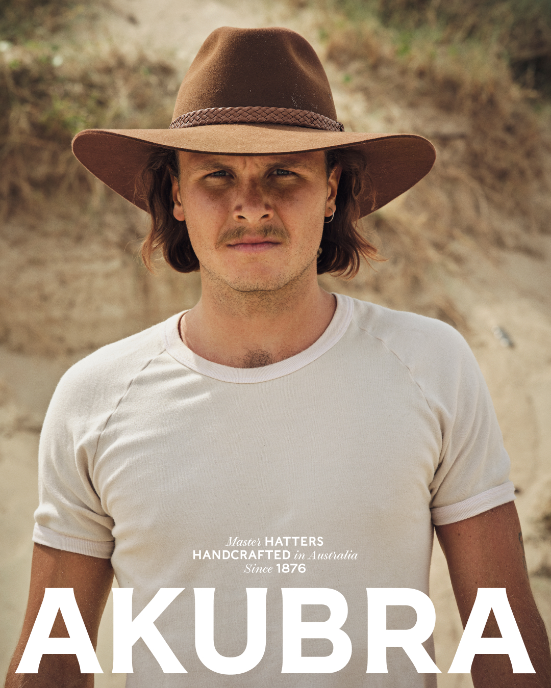 A person with long brown hair and a mustache wears a tan t-shirt and a brown hat in a close-up outdoor portrait on a sunny day.