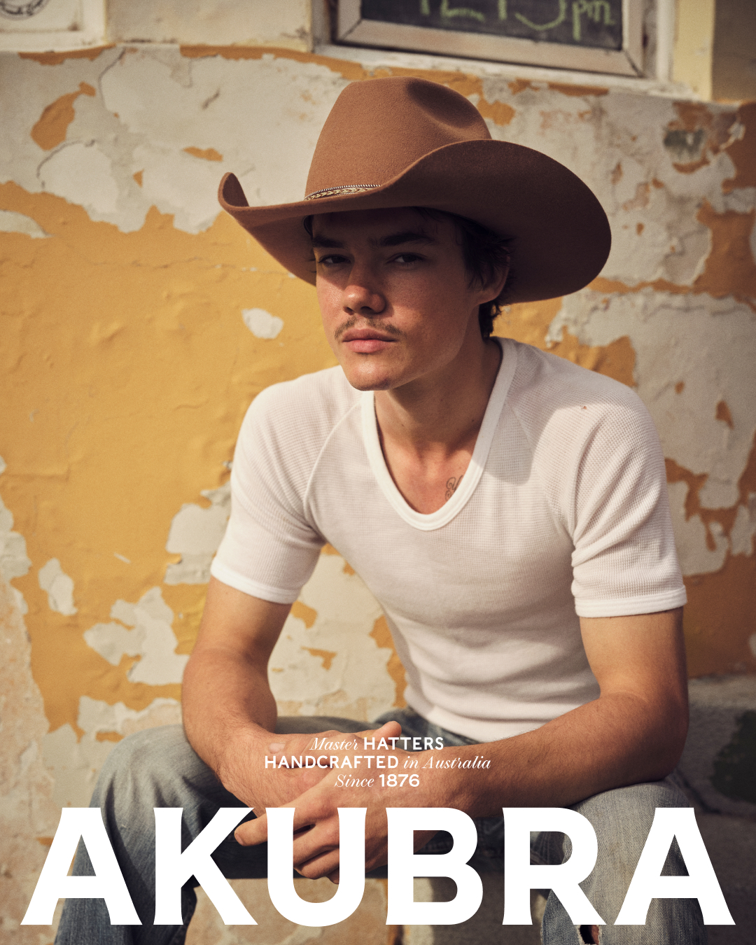 Man in a white t-shirt and jeans sits against an aged yellow wall wearing a brown Akubra cowboy hat for a fashion editorial.