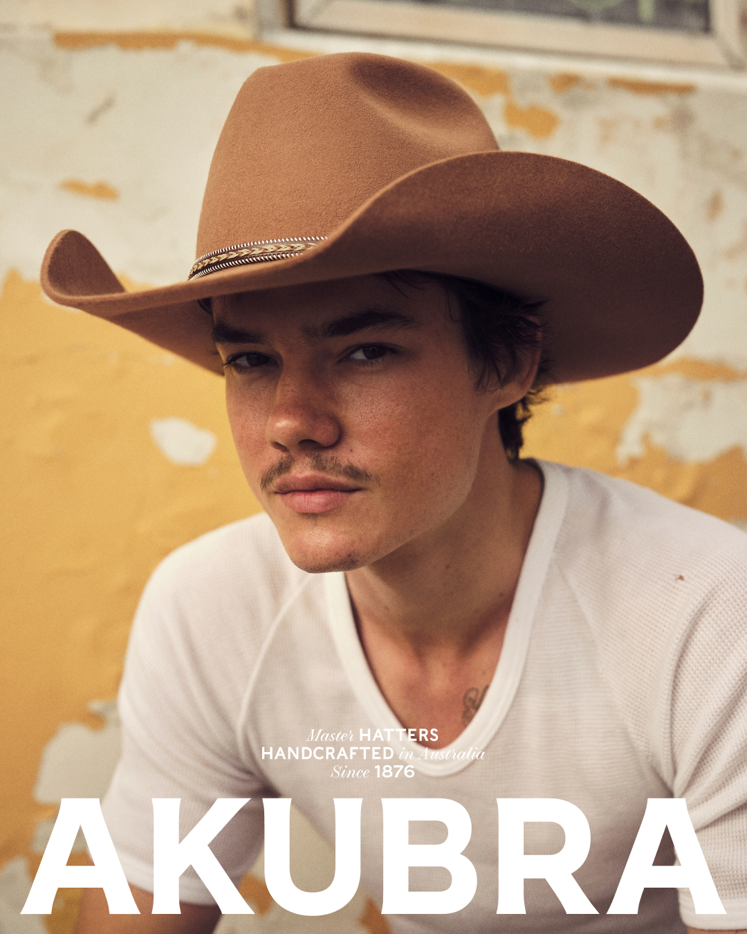 A young adult man wearing a cowboy hat, posing in front of a yellow wall for an advertisement, indoors in a studio with natural lighting.