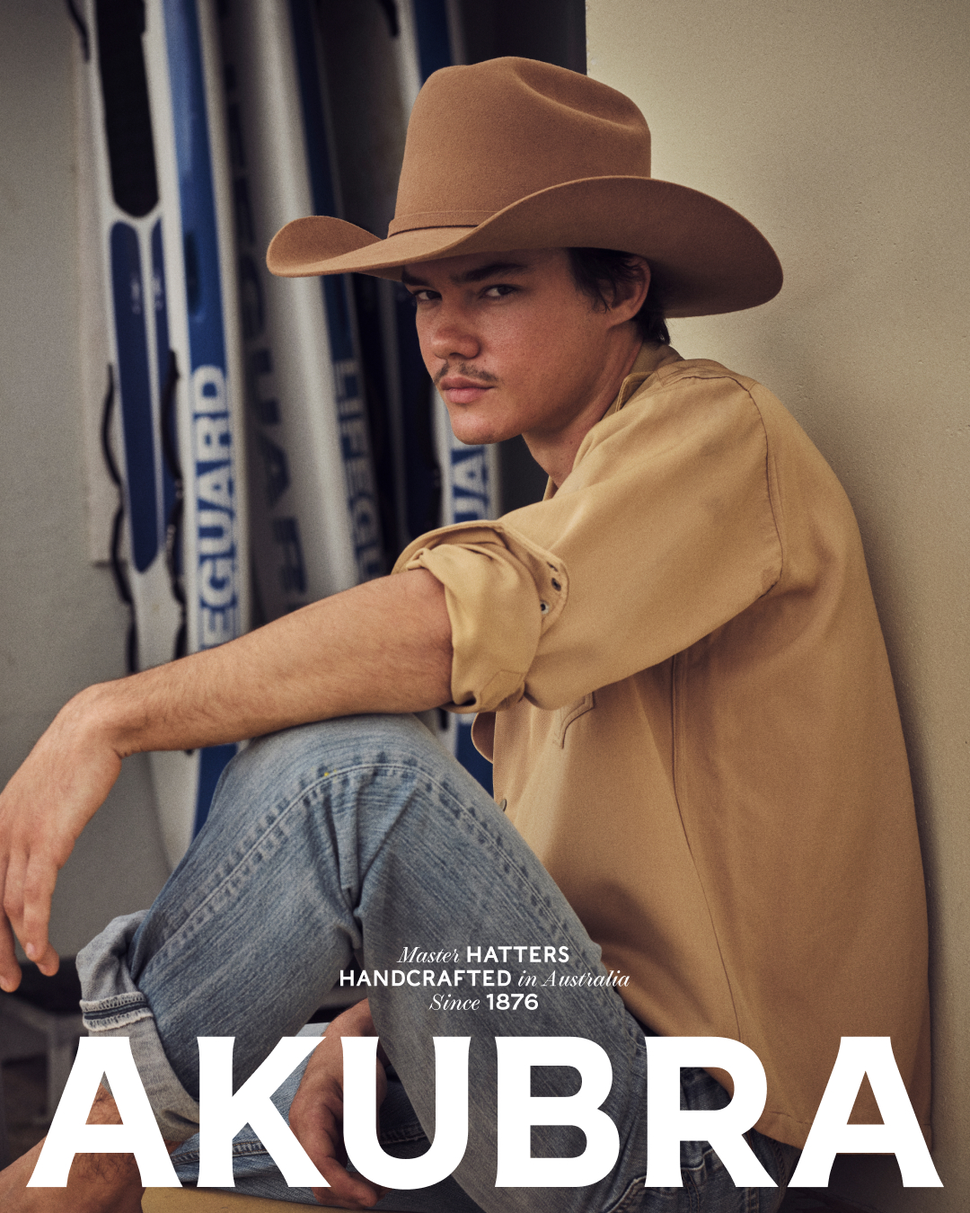 A man in a cowboy hat and denim jeans sits in front of surfboards.  He wears a tan long-sleeve shirt in an indoor setting.