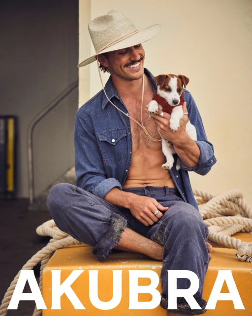 A bare-chested man in a cowboy hat sits holding a Jack Russell Terrier puppy with the text 'AKUBRA' in bold white on the bottom.