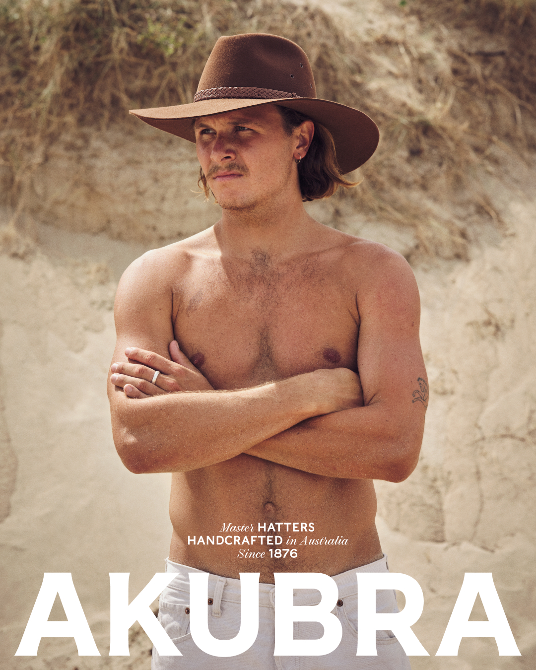 Shirtless male model poses with brown hat near tan dune. Fashion portrait with natural light, neutral background, and clear brand visibility.