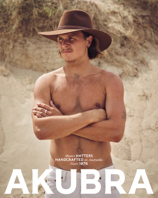 Shirtless male model poses with brown hat near tan dune. Fashion portrait with natural light, neutral background, and clear brand visibility.