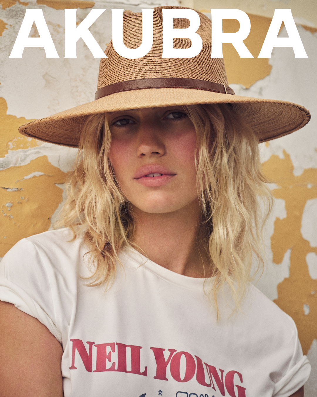 Woman wearing a hat poses against a textured wall. The image highlights a straw hat with a brown band and a white graphic tee.