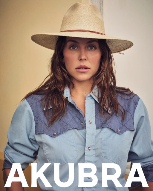 Portrait of a woman wearing a straw hat and blue denim shirt against a neutral background. She is posing for the camera.