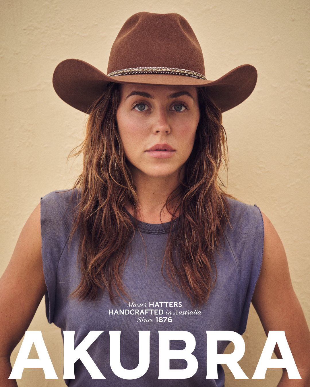Portrait of a woman wearing a brown cowboy hat and a sleeveless gray top in front of a neutral background with the brand AKUBRA.