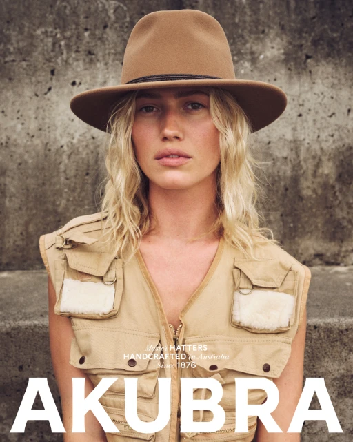 A woman with blonde hair, wearing a hat and utility vest, poses in front of a textured wall, looking directly at the camera.