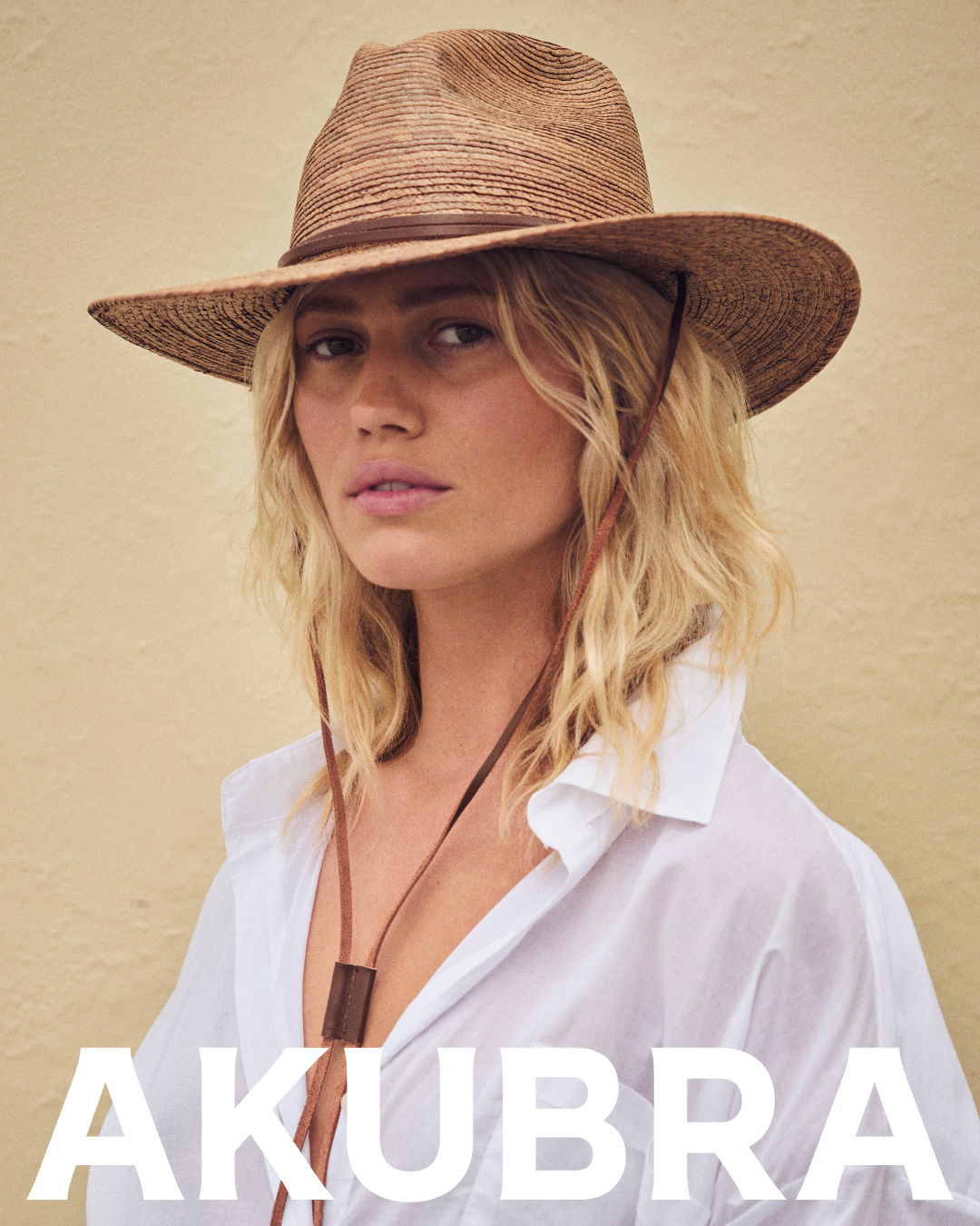 Close-up of a woman wearing a straw hat with a brown leather strap, and a white collared shirt against a beige background.