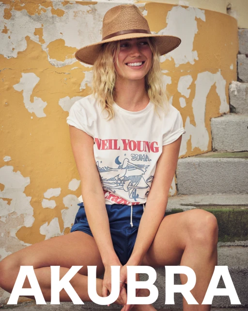Blonde woman in a Neil Young t-shirt and blue shorts wears a straw hat while sitting on stone steps with a yellow wall backdrop.