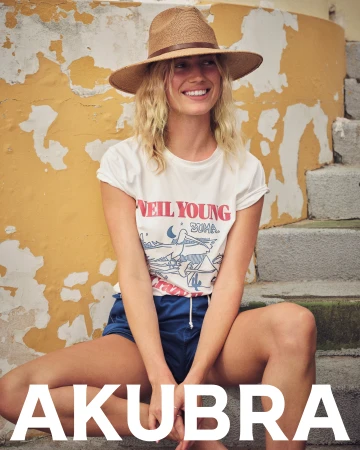 Blonde woman in a Neil Young t-shirt and blue shorts wears a straw hat while sitting on stone steps with a yellow wall backdrop.