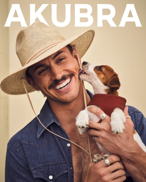 Smiling man in a straw hat holds a Jack Russell Terrier puppy licking his face, wearing a denim shirt and holding the dog closely.