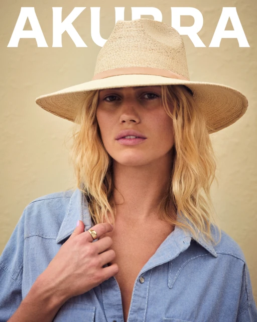 Woman with blonde wavy hair wearing a light denim shirt and straw hat against a light beige backdrop, portrait-style fashion photograph.