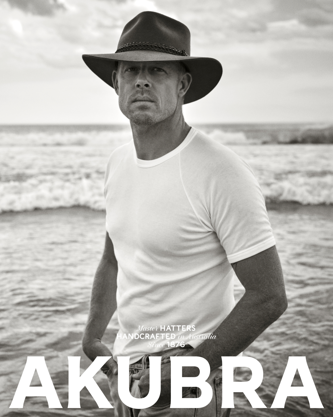 Monochrome portrait of a man wearing an Akubra hat standing on a beach with gentle waves; the brand name is prominently displayed.