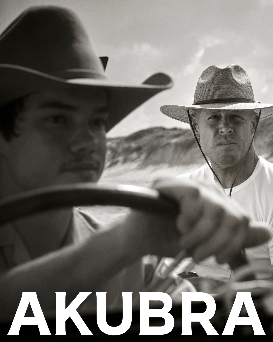 Black and white portrait of two men wearing Akubra hats in a vehicle, with a background of sand dunes under a bright sky.