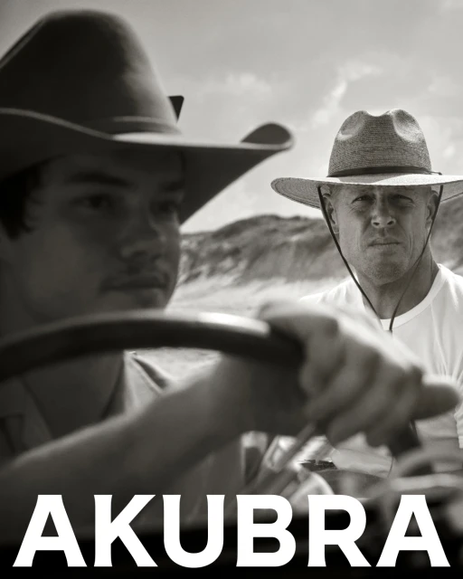 Black and white portrait of two men wearing Akubra hats in a vehicle, with a background of sand dunes under a bright sky.