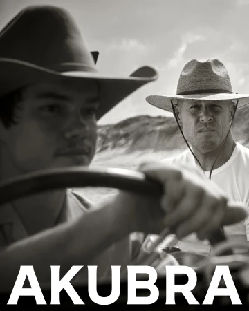 Black and white portrait of two men wearing Akubra hats in a vehicle, with a background of sand dunes under a bright sky.