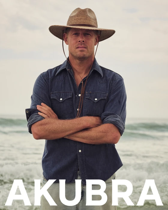 Mick Fanning posing on a beach with crossed arms, wearing a brown wide-brim Akubra hat and a blue denim western shirt.