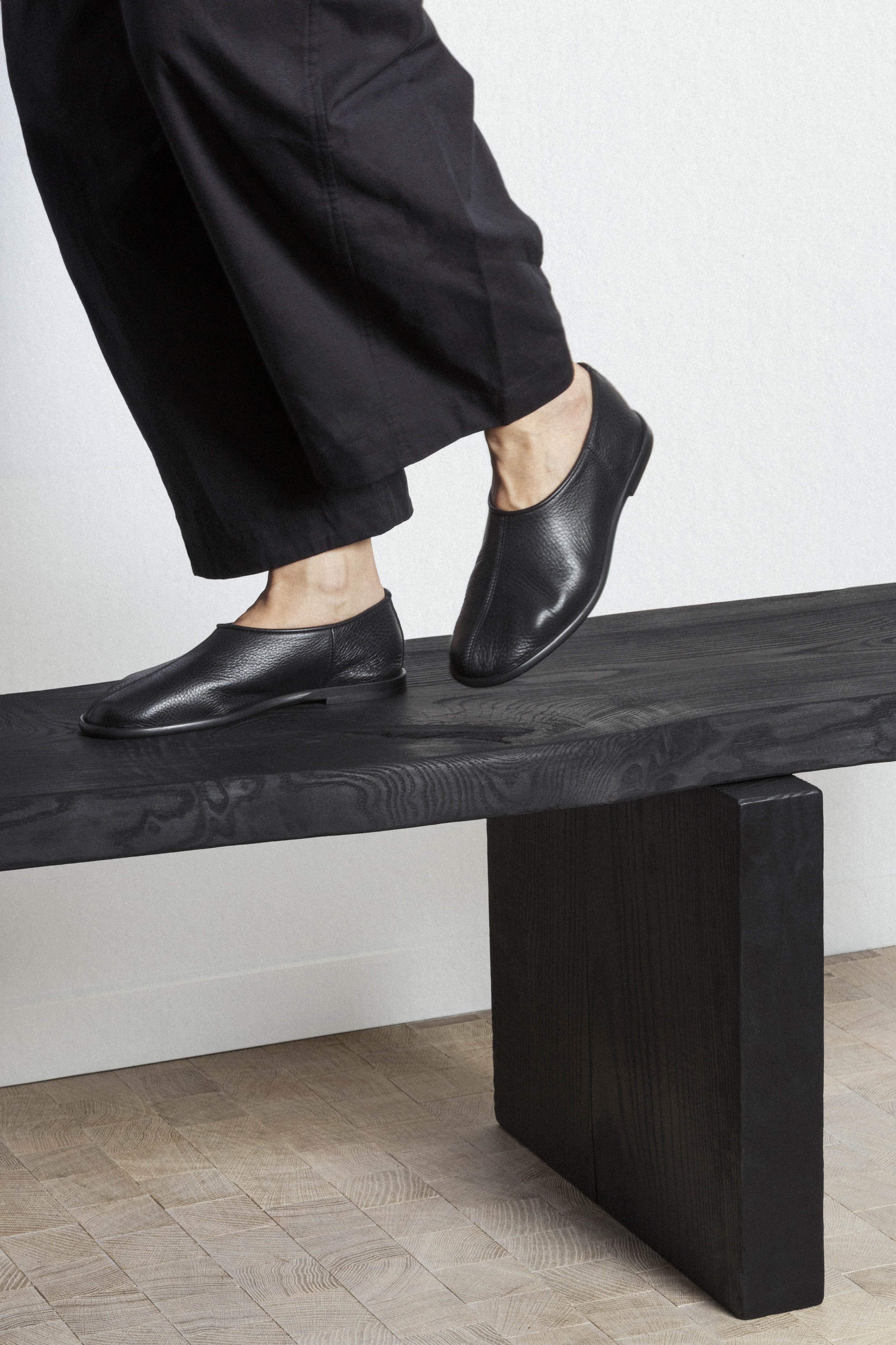 Black leather shoes on a black wooden bench, featuring a person in black pants about to step onto the bench. Minimalist footwear.