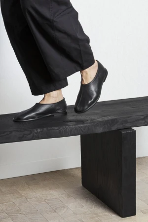 Black leather shoes on a black wooden bench, featuring a person in black pants about to step onto the bench. Minimalist footwear.