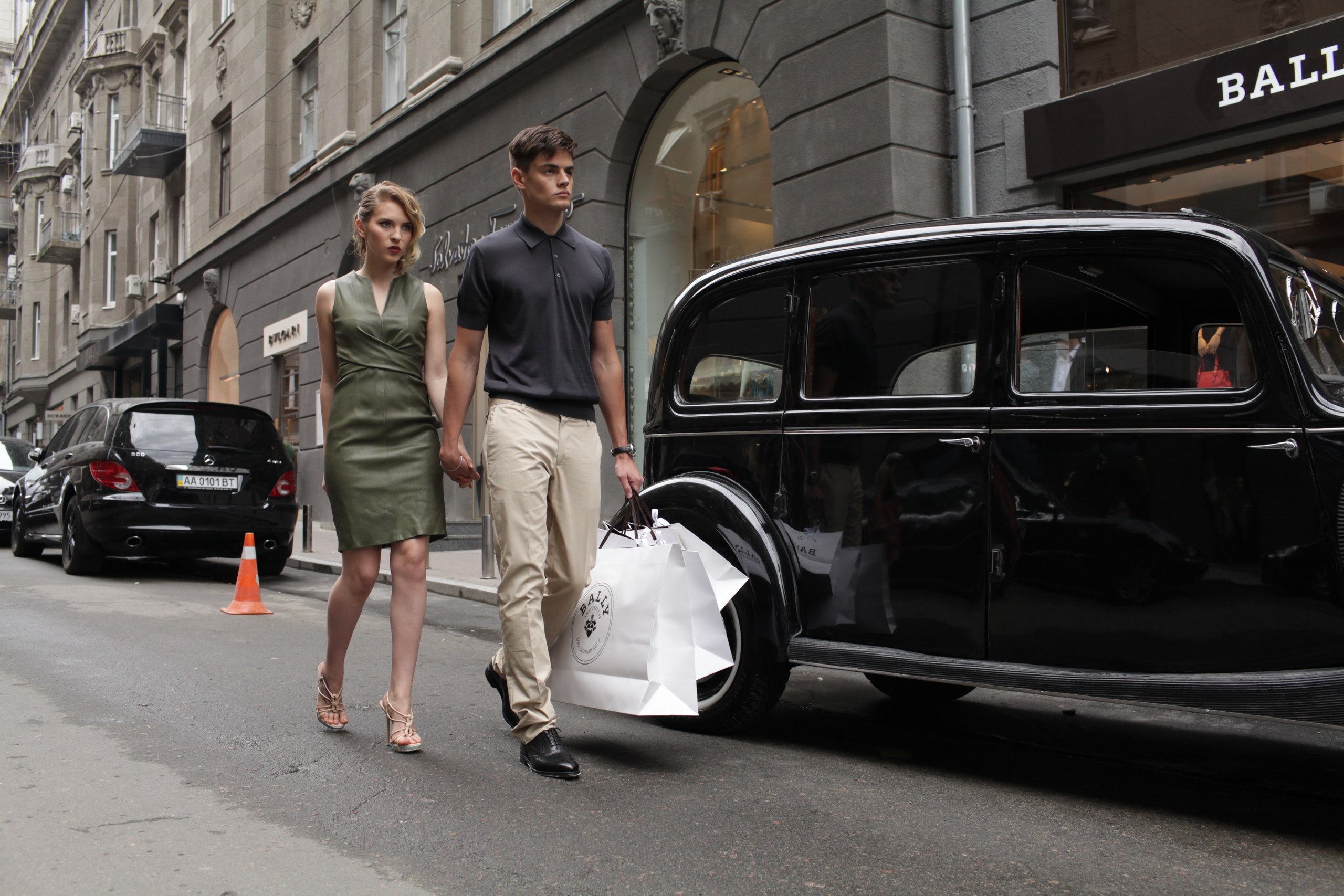 Fashionable couple strolls down city street with shopping bags, classic black car, and cityscape buildings in the background.