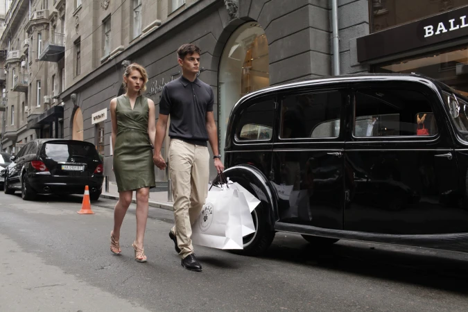 Fashionable couple strolls down city street with shopping bags, classic black car, and cityscape buildings in the background. 