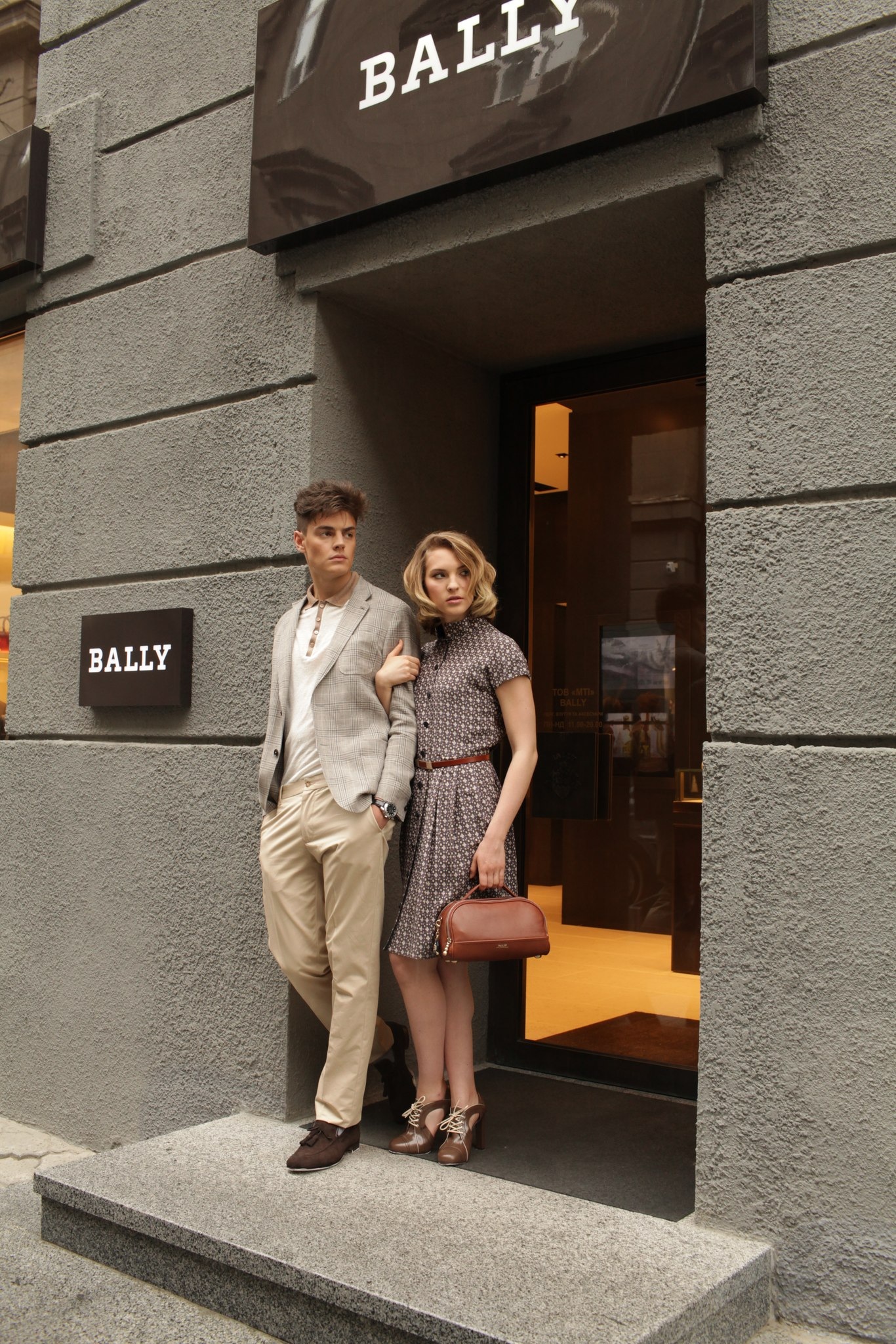 A fashionable couple poses outside a Bally store. The man wears a blazer and khaki pants, while the woman dons a polka-dot dress with vintage-style shoes.
