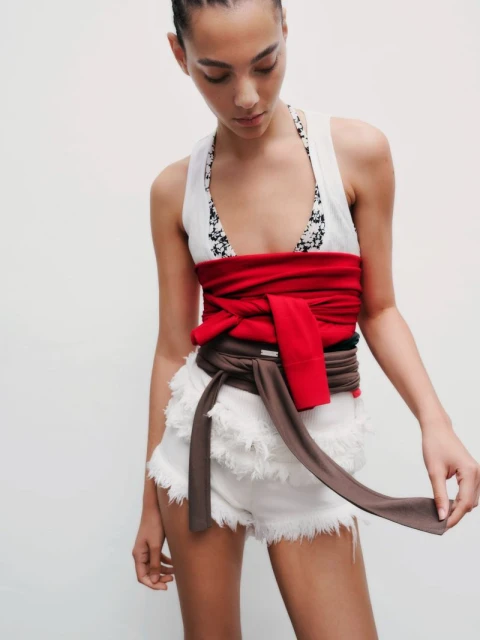 Young woman in a halter top and white frayed denim shorts, wearing a red and brown wrap belt, posing against a white background.