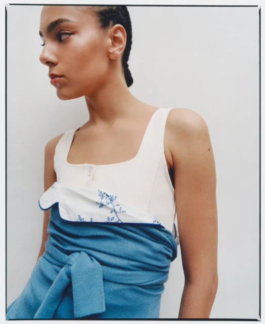 Young woman poses in an off-white top featuring floral embroidery and a blue woven wrap against a neutral background.