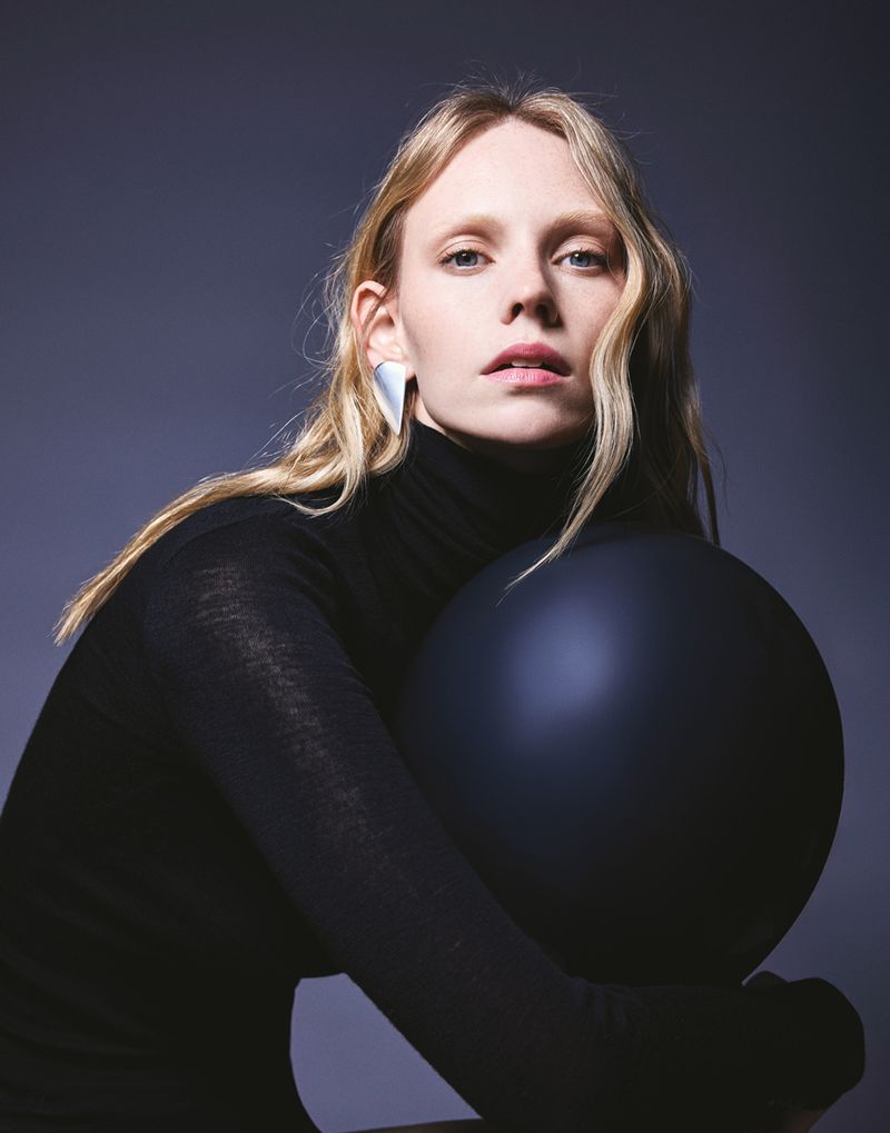 Blonde woman with fair skin and sleek black turtleneck embraces a smooth, dark blue sphere in a dimly lit indoor setting.