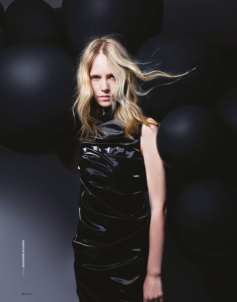 Portrait of a fair-haired woman in a black patent dress surrounded by black balloons against a dark grey gradient background in a studio.