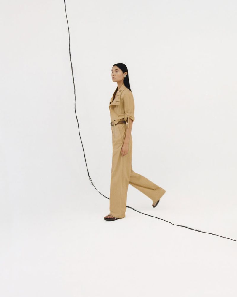 A side-view shot of an Asian woman in a tan jumpsuit walking against a stark white backdrop with a black cable in the foreground.