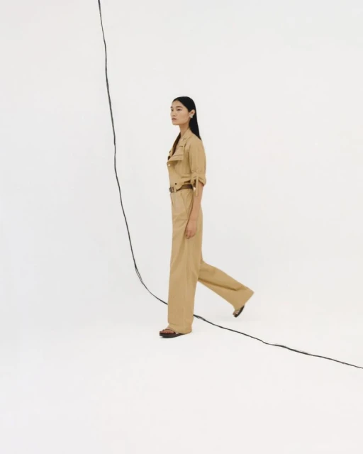 A side-view shot of an Asian woman in a tan jumpsuit walking against a stark white backdrop with a black cable in the foreground.